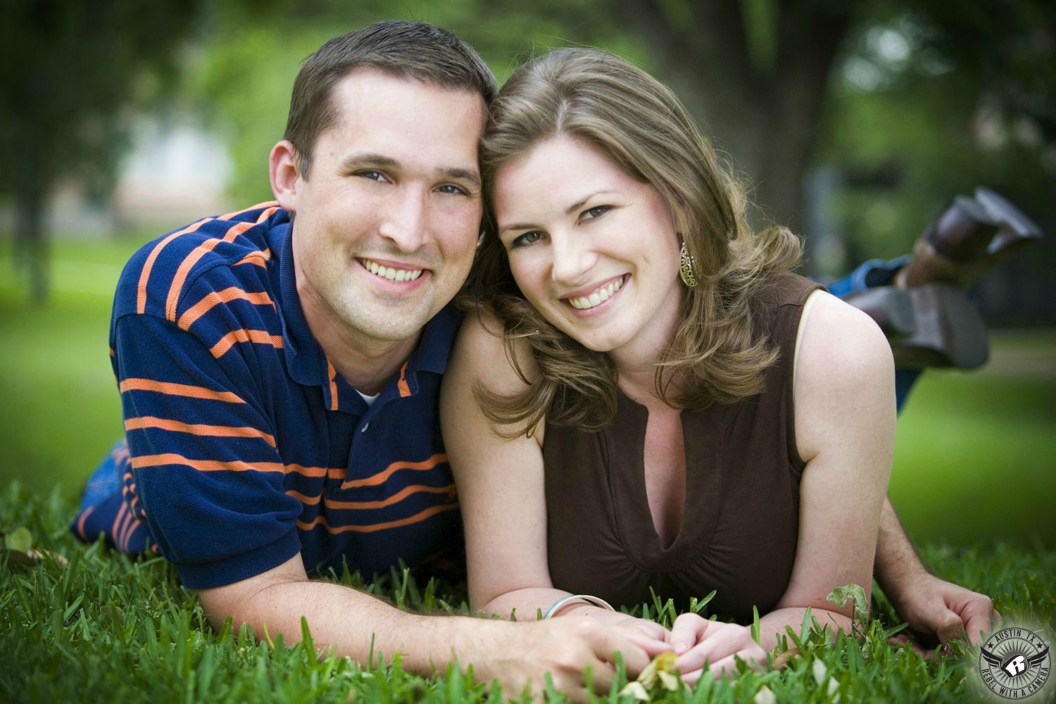 engagement couple happily lying in grass up on their elbows with blue and orange striped shirt and brown low cut blouse with brown hair at the texas capital grounds 